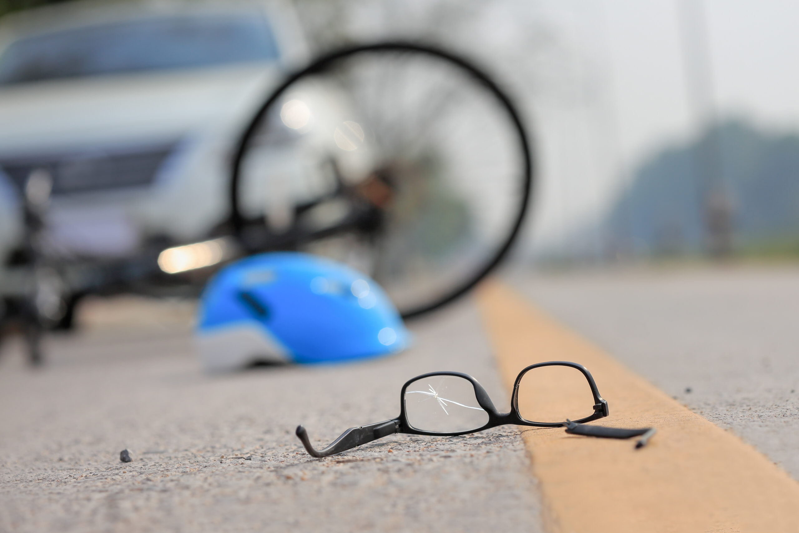 broken glasses lay on the road and a bicycle and bicycle helmet rest on the road with a car parked behind them signifying a car and bike accident